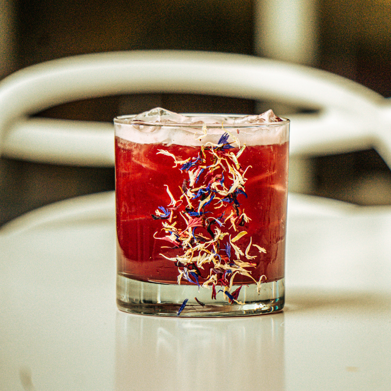deep red bourbon cocktail in a glass filled with ice. Glass is garnished with cornflower fireworks 