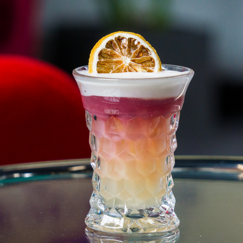 bourbon cocktail with a light yellow colour at the bottom, purple colour top and a white egg white foam on the top, with a dried lemon garnish in a glass. 