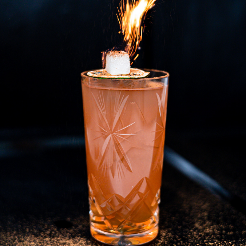 All-Inclusive Cocktail - spiced rum, grapefruit, guava, cinnamon, orgeat, falerum, lime, absinthe in a collins glass garnished with lime and sugar cube on fire from  absinthe  and cinnamon 